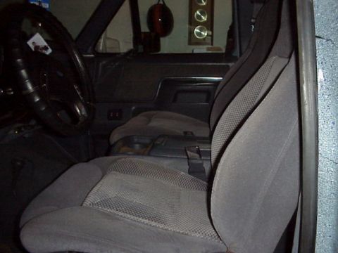 1989 Ford Bronco - Front cab of a Ford truck viewed through the open passenger door, showing gray cloth high-back bucket seats with a seat belt draped across…