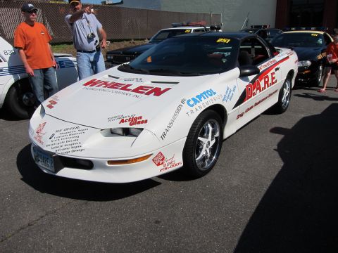 1991 Ford LTD Crown Victoria - white Chevrolet Camaro covered in Whelen, Capitol, and D.A.R.E. decals parked among police cars with onlookers nearby.