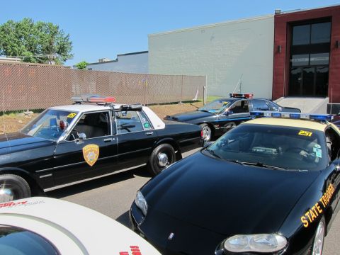 1991 Ford LTD Crown Victoria - black-and-white police sedan beside a black 'State Trooper' Chevrolet Camaro parked along a fence by a building.