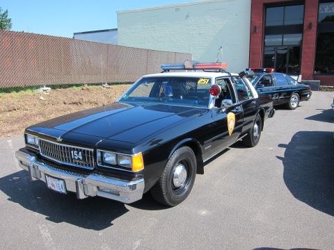 1991 Ford LTD Crown Victoria - black Chevrolet Caprice police sedan numbered 154 with a roof lightbar parked in a lot beside another black cruiser.