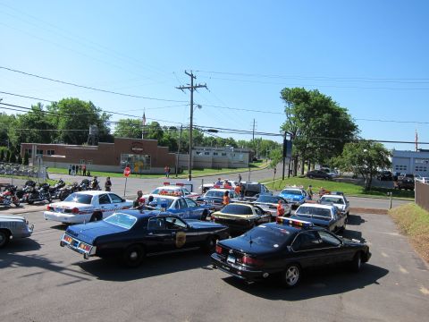 1991 Ford LTD Crown Victoria - Elevated view of black, white, and silver police cars plus motorcycles and onlookers filling a paved lot at a gathering.