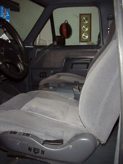 1989 Ford Bronco - Interior of a Ford truck shot through the open passenger door, showing a gray cloth bucket seat with a plastic power-seat base and switches…