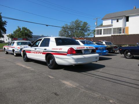 1991 Ford LTD Crown Victoria - Two white Cayuga County Sheriff Ford Crown Victoria sedans parked on pavement near a house, with other classic cars behind.