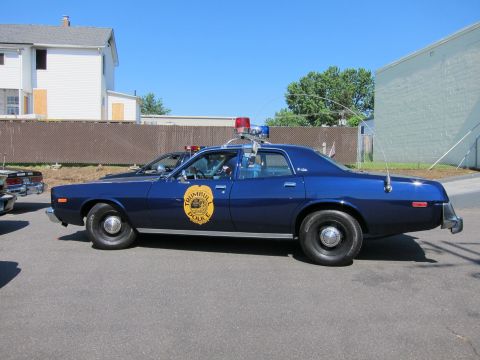 1991 Ford LTD Crown Victoria - dark blue vintage Plymouth Trumbull Police sedan with a gold door seal and roof lights parked along a fence by houses.