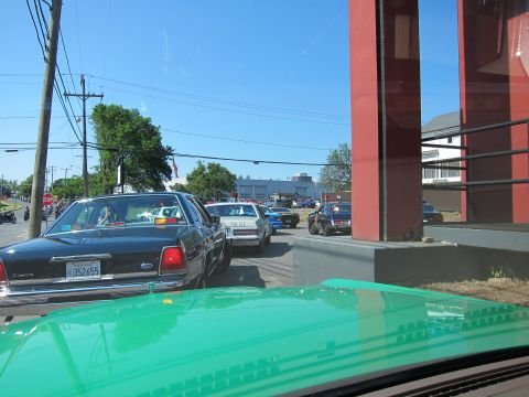 1991 Ford LTD Crown Victoria - Looking over a green car hood at a queue of cars and police cruisers along a sunny street near a red-pillared building.