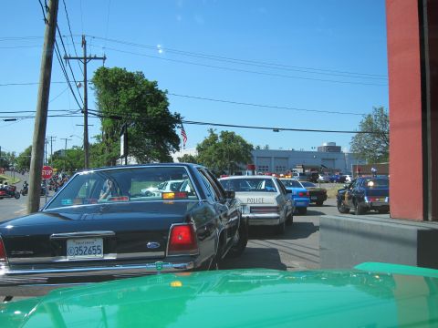 1991 Ford LTD Crown Victoria - black Ford sedan leads a line of police cruisers parked near a building under a clear blue sky.
