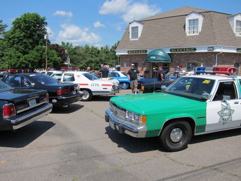 1991 Ford LTD Crown Victoria - green-and-white sheriff car sits among classic police cruisers in a parking lot outside Modugno Electric.