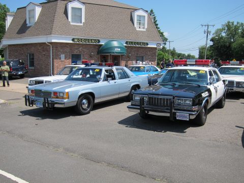 1991 Ford LTD Crown Victoria - silver police sedan and a black-and-white police cruiser parked outside a brick building at a show.