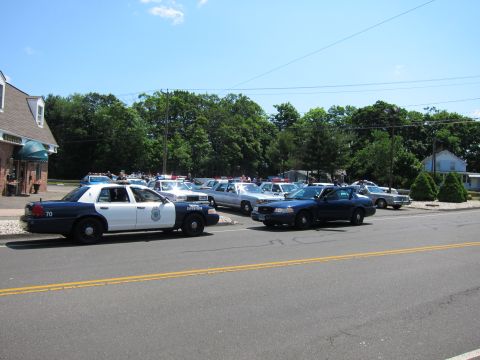 1991 Ford LTD Crown Victoria - Police cruiser marked 70 and a lot full of parked police cars under a bright blue sky at a car show.