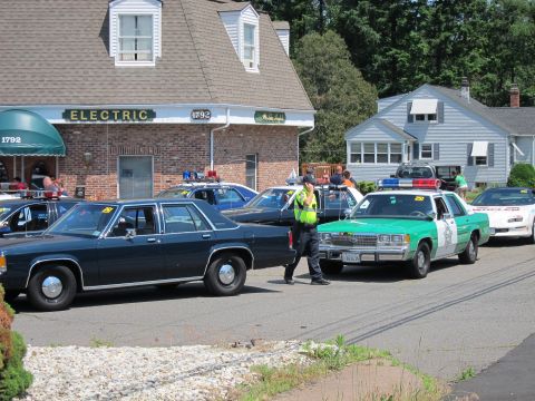 1991 Ford LTD Crown Victoria - officer in a safety vest directs a green police car past a row of parked cruisers outside a building.