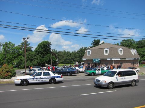 1991 Ford LTD Crown Victoria - Police cruisers and classic cars line a brick building's lot while a white minivan passes on the road in front.