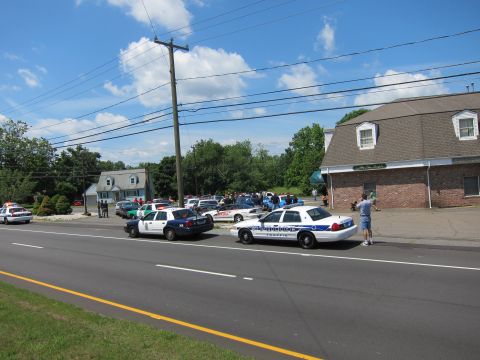 1991 Ford LTD Crown Victoria - Police cruisers parked at angles in a lot beside the road near a brick building, with onlookers nearby.