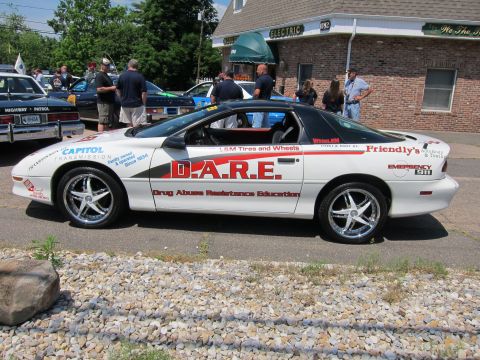 1991 Ford LTD Crown Victoria - white Chevrolet Camaro D.A.R.E. car covered in sponsor decals, parked on gravel with people and cars behind.