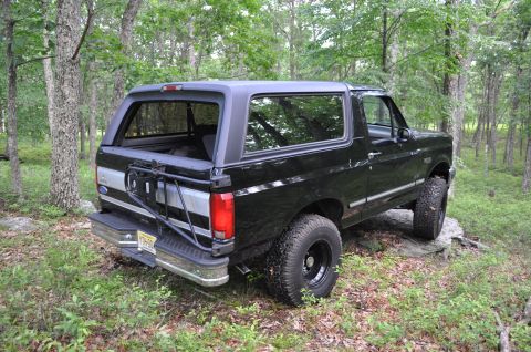 1995 Ford Bronco - This black 1995 Ford Bronco is parked off-road in a wooded area, showing its rear and side profile.