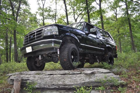 1995 Ford Bronco - A black Ford Bronco is positioned on large rocks in a wooded area, with trees surrounding it.