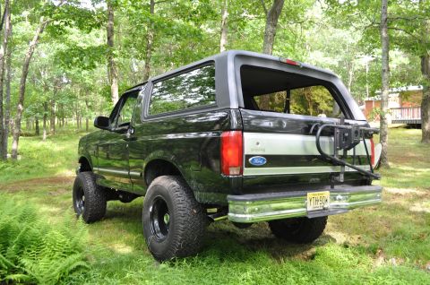 1995 Ford Bronco - Rear view of a black 1995 Ford Bronco XLT parked in a grassy, wooded area. It has a spare tire carrier.