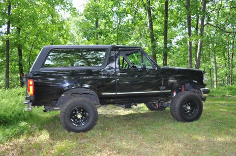 1995 Ford Bronco - This black 1995 Ford Bronco, seen from the side, features a lifted suspension and off-road tires, parked amidst a lush green forest.