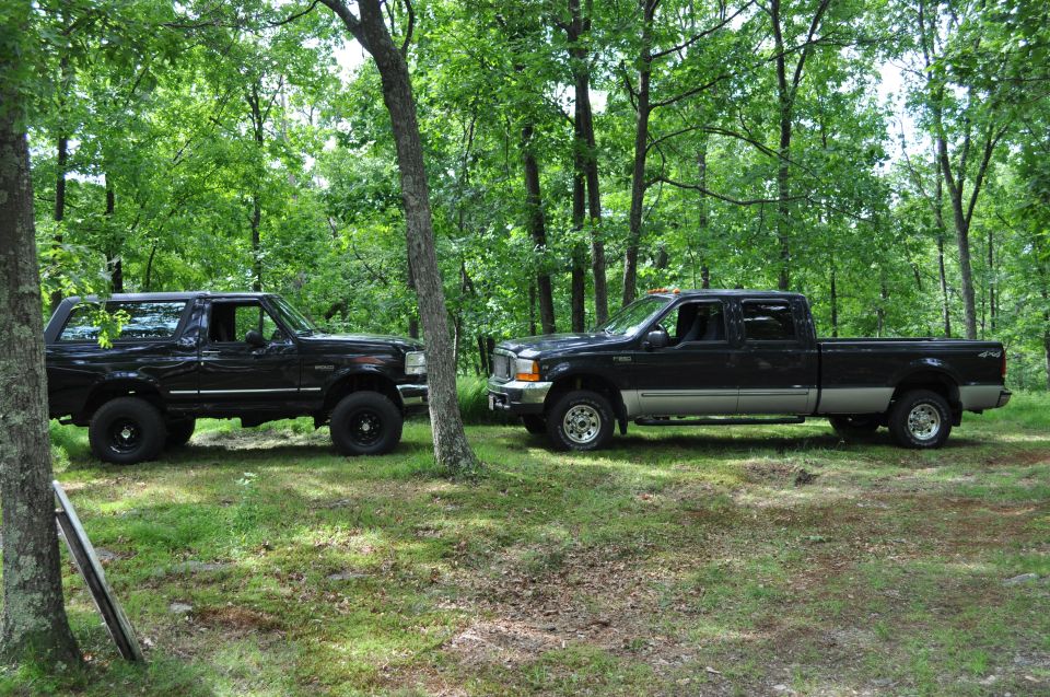 Black 1995 Ford Bronco and a black Ford F-250 Super Duty crew cab parked side-by-side in a forest clearing