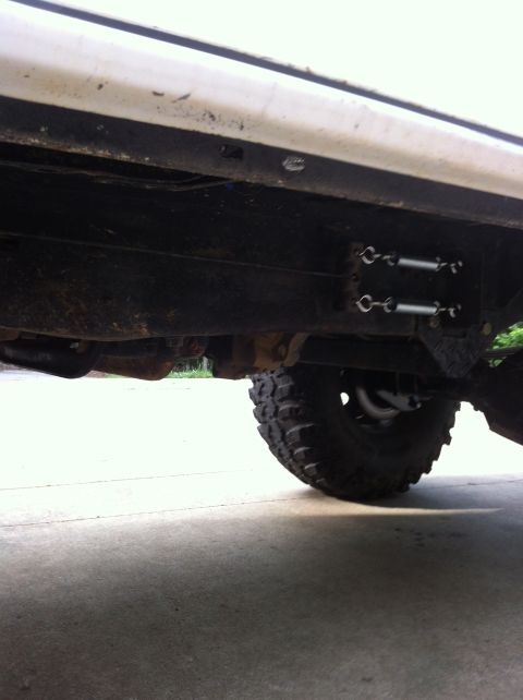 1996 Ford Bronco - Low ground-level view under the rocker of a white lifted 4x4, showing the rusty frame rail, skid/crossmember plate, and two turnbuckle-styl…