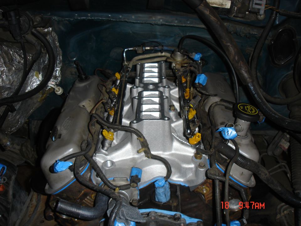 Engine bay with a shiny intake manifold, yellow fuel injectors, and various hoses and wires. Some parts are covered with blue tape