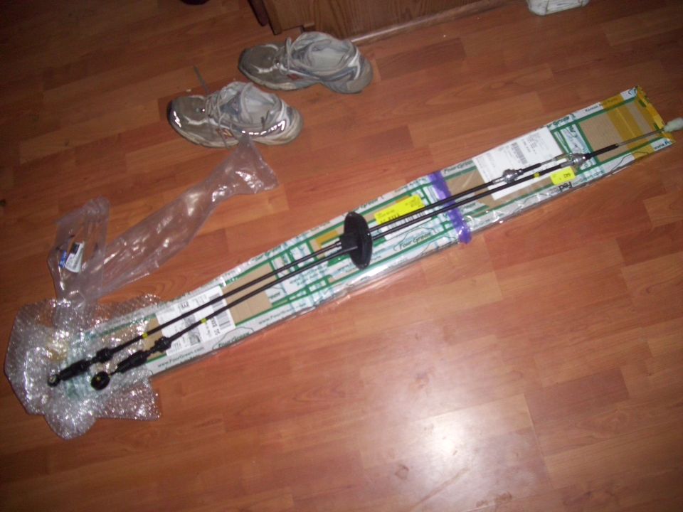 Two black control cables in packaging on a wood floor, with a pair of sneakers in the background
