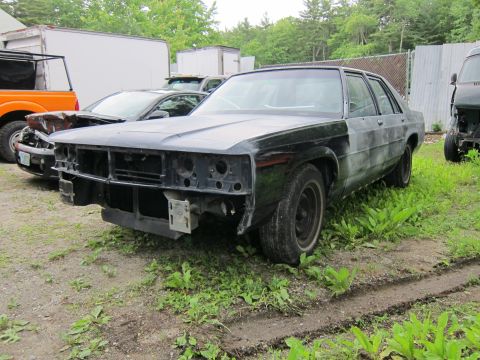 1991 Ford LTD Crown Victoria - A dark sedan sits in a yard, its front end dismantled, with other vehicles visible in the background.