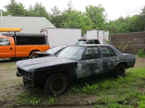 1991 Ford LTD Crown Victoria - A dark sedan, possibly a Ford Crown Victoria, sits in a grassy lot with other vehicles and trailers visible in the background.
