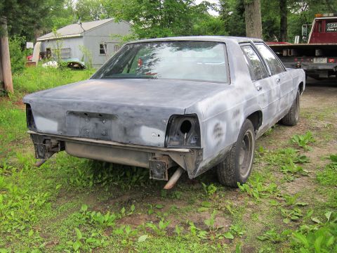 1991 Ford LTD Crown Victoria - This sedan, covered in primer and missing its taillights, sits in a grassy area next to a tow truck.