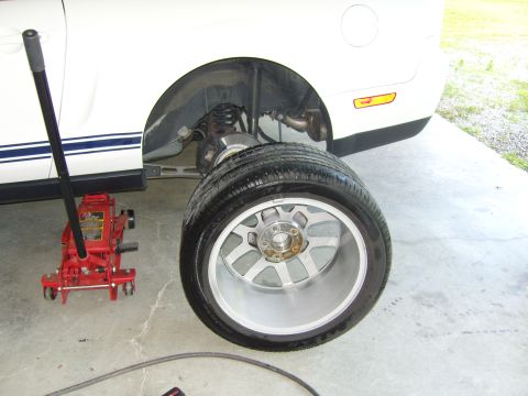 2007 Ford Mustang - A white Ford Mustang with blue racing stripes is shown with its rear wheel removed and a red jack underneath.