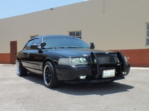 2003 Ford Crown Victoria - Front 3/4 in lot; black sedan on black wheels with whitewalls, push bar, EVIL VIC plate