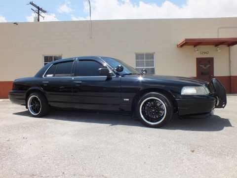2003 Ford Crown Victoria - Right profile in lot; long black sedan, push bar, building door numbered 12942 behind