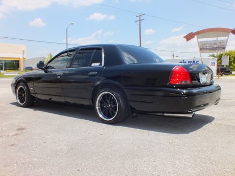 2003 Ford Crown Victoria - Left rear 3/4; long black sedan, tinted windows, industrial storefronts and sign behind
