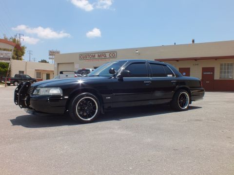 2003 Ford Crown Victoria - Black sedan with grille guard and tinted windows angled toward the camera in a lot.