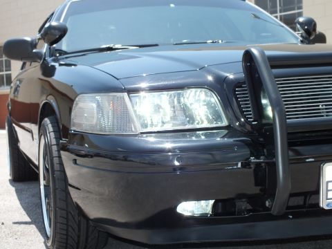 2003 Ford Crown Victoria - Detail of a black sedan's headlight, push bar and black multi-spoke wheel.