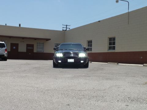 2003 Ford Crown Victoria - Head-on shot of a black sedan with headlights on near a tan building and van.