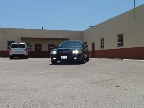 2003 Ford Crown Victoria - Black sedan with headlights on beside a silver SUV in a wide asphalt lot.