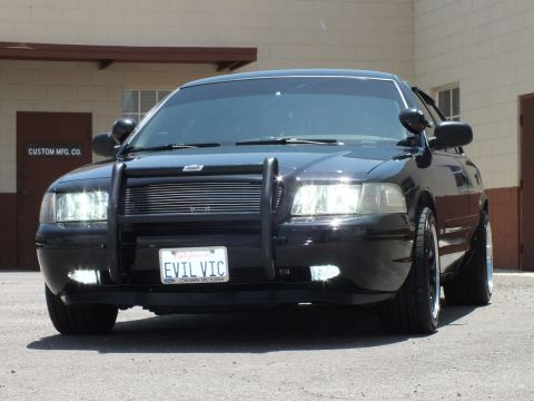 2003 Ford Crown Victoria - Lowered black sedan angled toward camera, billet grille and push bar in sunlight.