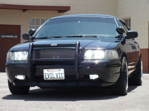 2003 Ford Crown Victoria - Sunlit black sedan front-quarter with brush guard and California vanity plate.