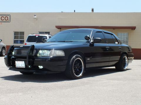 2003 Ford Crown Victoria - Driver-side profile of a lowered black sedan on dark polished-lip wheels by a warehouse.