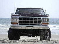 1986 Ford Bronco - A dark red Ford Bronco is parked on a sandy beach with the ocean in the background.