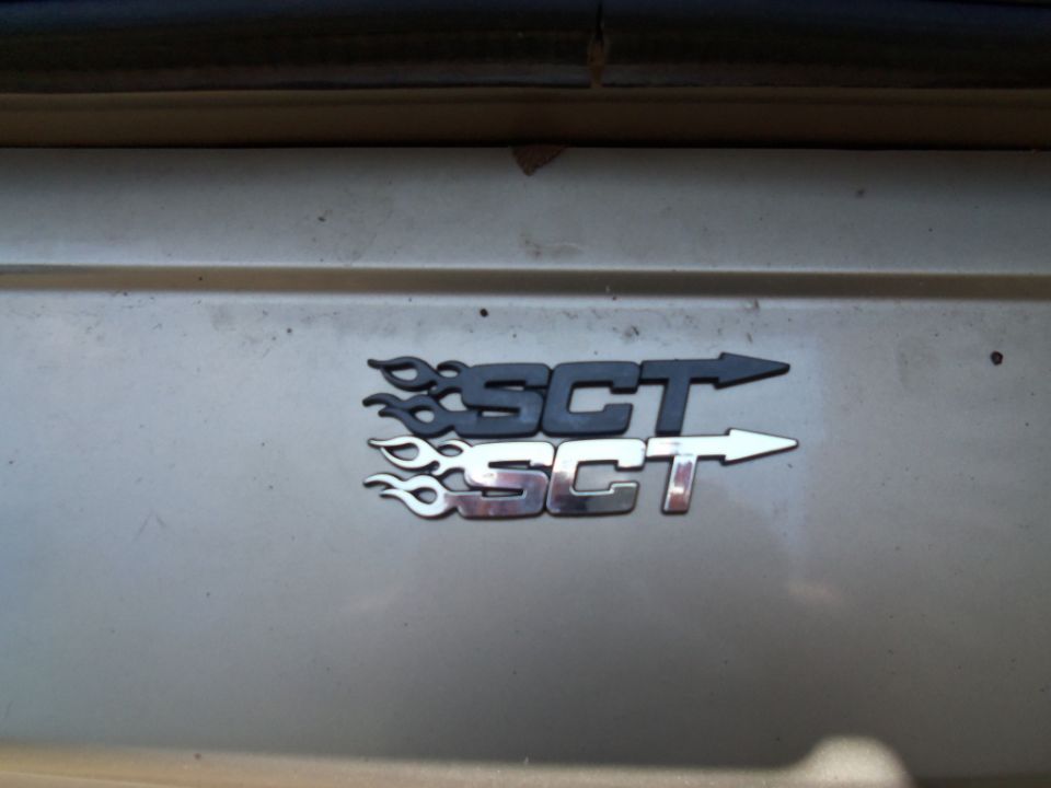 Two SCT emblems with flame graphics, one black and one chrome, on a car's body panel