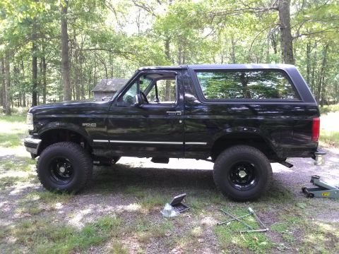 1995 Ford Bronco - A black Ford Bronco is parked on a dirt surface with trees in the background. Tools and a jack are on the ground next to it.