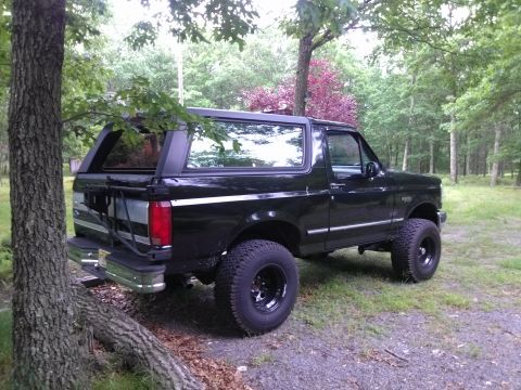 1995 Ford Bronco - A black Ford Bronco SUV is parked on a dirt path surrounded by trees.