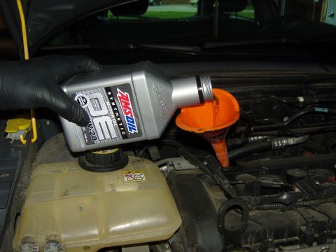 2007 Ford Mustang - A person pours AMSOIL synthetic motor oil into a vehicle's engine bay using an orange funnel.