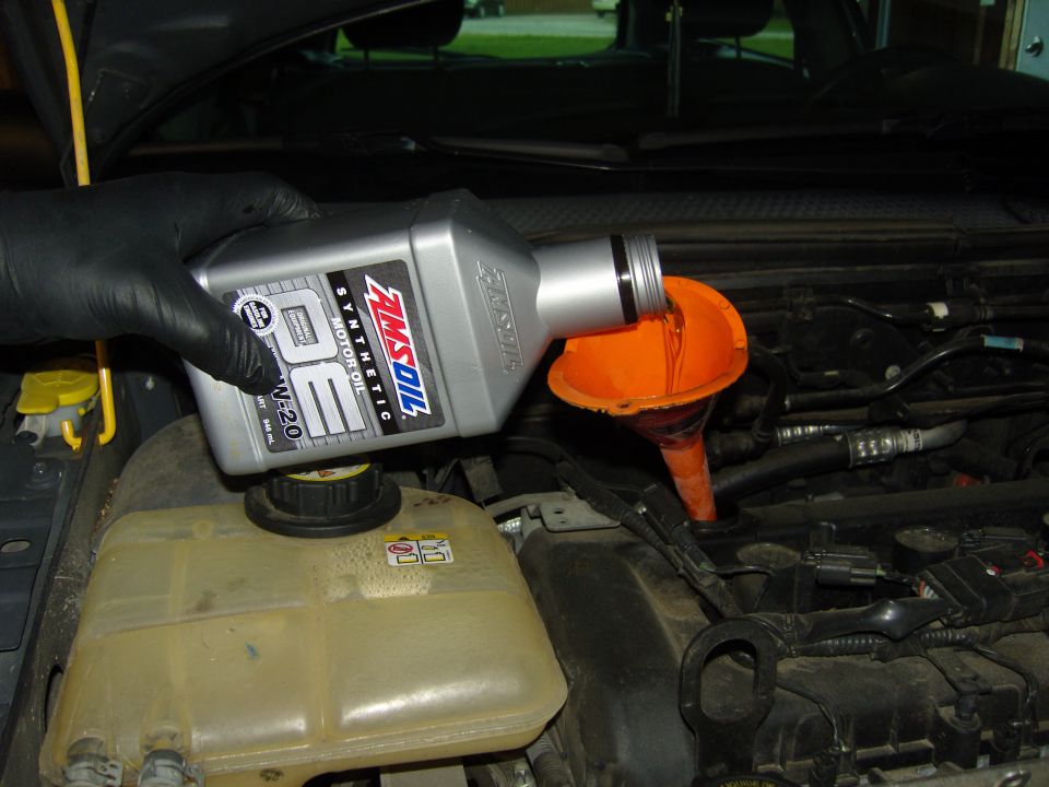 Engine bay with a gloved hand pouring AMSOIL synthetic motor oil into an orange funnel