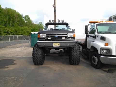 1995 Ford Bronco - A heavily modified Ford truck with oversized tires is parked next to a tow truck, its lifted suspension and front-end details.