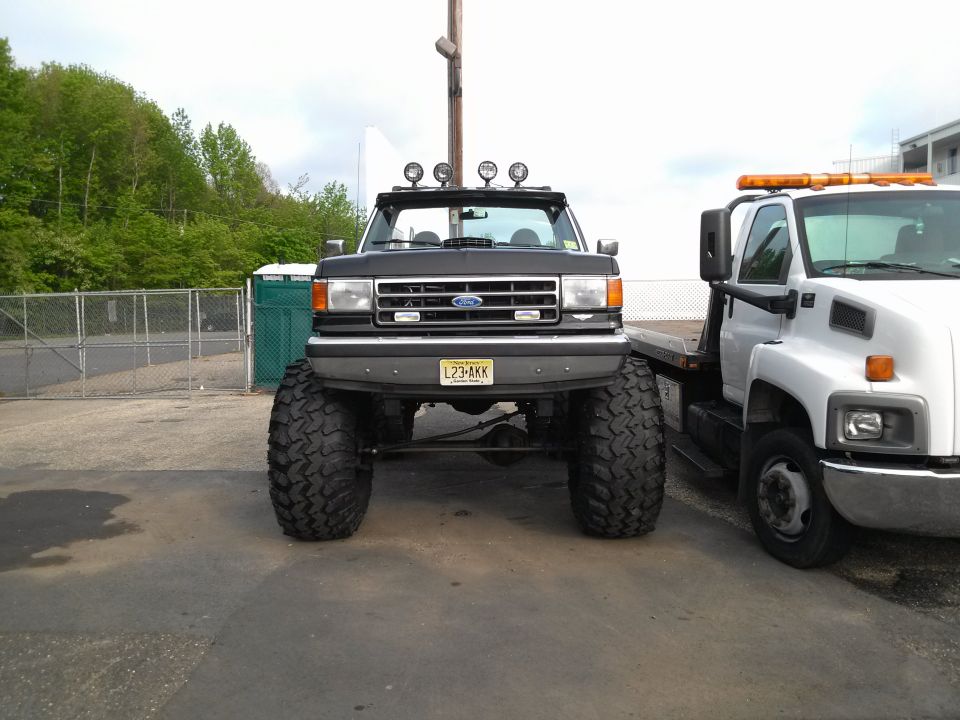 Front view of a lifted dark-colored Ford truck with large tires, parked next to a white tow truck. The truck has four auxiliary lights on the roof