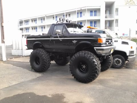 1995 Ford Bronco - Modified black Ford Bronco with large off-road tires sits beside a white utility truck.