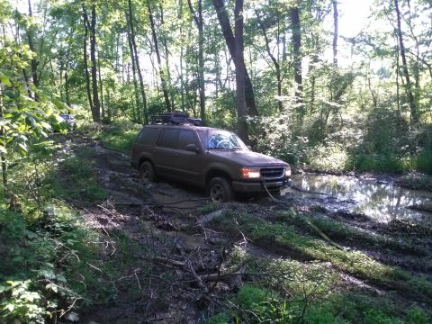 1995 Ford Bronco - A brown Ford Explorer navigates a muddy, wooded path, with another vehicle visible in the distance.