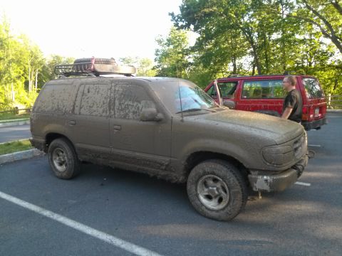 1995 Ford Bronco - Two SUVs are parked in a lot, one is heavily covered in mud and has a roof rack.
